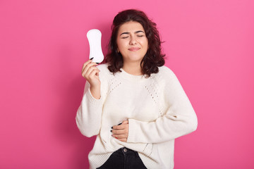 Wall Mural - Gloomy Caucasian woman cares about personal hygiene, holds sanitary pad in her hands, suffers from pain during menstruation, has unset facial expression, wears white outfit, poses in studio.