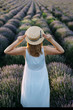 A girl in a white dress and straw hat looks at the lavender field, looking from the back