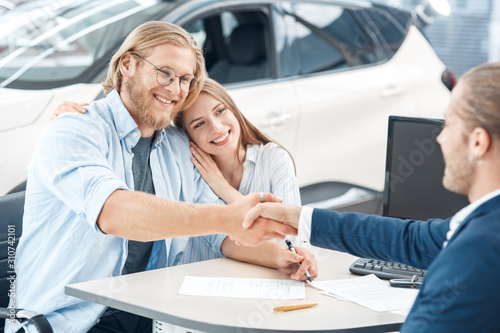 Car Salon. Young couple sitting at table man shaking hands with agent buying car cheerful