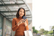 © makistock - Portrait of elegant young Asian woman holding shopping bags and using smartphone on the go while leaving mall