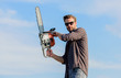 © be free - Sharp blade. Handsome man with chainsaw blue sky background. Gardener lumberjack equipment. Lumberjack with chainsaw in his hands. Dangerous job. Feeling confident. Risky temper. Powerful chainsaw