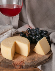 Wall Mural - Caciotta cheese on a round wooden board. Next to a sliced ​​piece of cheese, black grapes and a glass of red wine. Background gray fabric from linen.