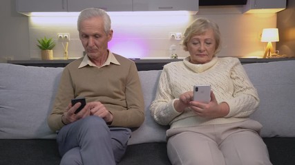Modern elderly man and woman scrolling phone app, sitting on sofa, internet