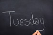 Woman's hand writing the day of the week on a blackboard with white chalk, tuesday