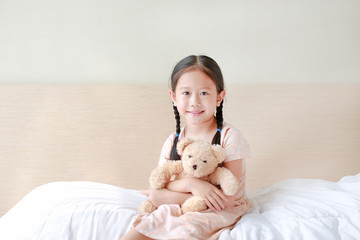 Wall Mural - Smiling Asian little girl cuddle teddy bear and looking camera while sitting on the bed at home.