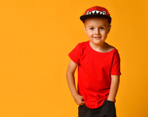 Young guy boy in a red T-shirt and dark pants, white sneakers and a funny cap posing on a free copy space on a yellow background