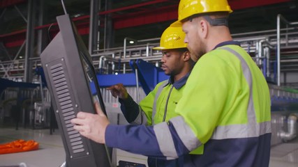 Two diverse male engineers in uniform and hard hats use computer LCD touch screen for programming CNC machine. Work process on heavy industry plant or factory. Employees and high tech