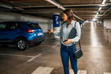 Woman In A Parking Garage, Unlocking In Her Car. Woman Activating Her Car Alarm In An Underground Garage As She Walks Away. Young Business Woman Walking With Car Keys In The Underground Parking.