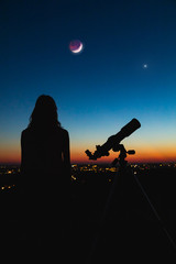 Wall Mural - astronomer with a telescope watching at the stars and moon with blurred city lights in the backgroun