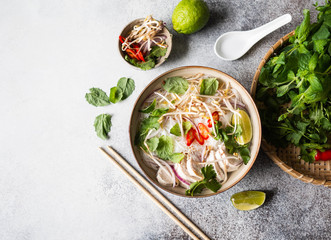 Traditional Vietnamese soup- pho ga in bowl with chicken and rice noodles, mint and cilantro, red onion, chili, bean sprouts and lime on grey background. Asian food. Copy space. Top view