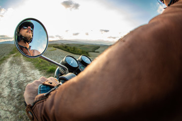 Reflection on rearview mirror of man riding motorbike along a country road on hills landscape. Tuscany, Italy.