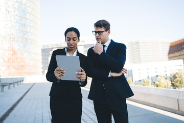 Collaborating man and woman using tablet Confident young businessman in glasses watching tablet with serious African American coworker having professional discussion on background of city buildings