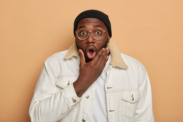 Wall Mural - Portrait of shocked black hipster guy opens mouth, holds breath from surprisement, wears white stylish clothing, optical glasses, feels impressed and very excited, isolated on brown background