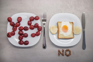 baked egg inside toast bread with two round croutons on a white plate with the inscription NO from grains below and the inscription YES from grapes on a white plate on a gray kitchen table top view