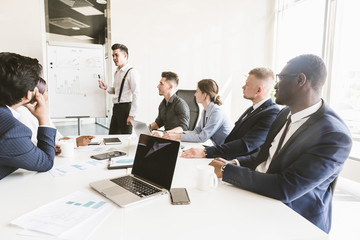 Wall Mural - Company leader standing near the flipchart. A team of young businessmen working and communicating together in an office. Corporate businessteam and manager in a meeting