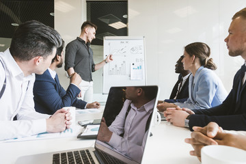 Wall Mural - Company leader standing near the flipchart. A team of young businessmen working and communicating together in an office. Corporate businessteam and manager in a meeting