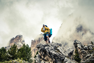 Wall Mural - young man hiker taking pictures on stone mountain with cloudy sky and fog. yellow jacket, backpack, 
