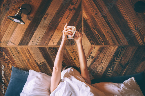 Lazy female lying under the white blanket on the  linen bed and holding the fresh coffee cup in the early morning. Lazy day off and \