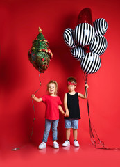 Wall Mural - Full length shot of little girl and boy in stylish clothes, looking at camera, smiling, holding colorful balloons, posing in red studio. Kids fashion