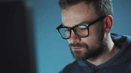 Male hacker in glasses working on a computer in a dark office room. Cybercrime concept