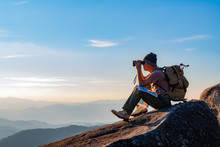 A Young Man With A Lifted Backpack Looked In Binoculars Sitting On Top Of The Mountain Enjoying A Spectacular View On The Mountains Above The Clouds.