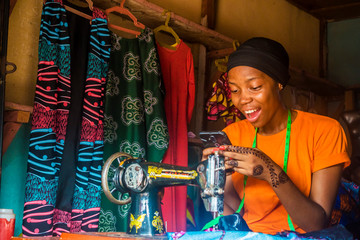 Poster - young african female tailor smiling while viewing content on her mobile phone sitting with her sewing machine