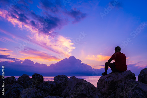 Cuadros en Lienzo Young man sitting on sea rocks by the beach thinking, contemplating, determining the way forward