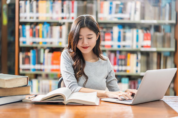 Wall Mural - Asian young Student in casual suit doing homework and using technology laptop in library of university or colleage with various book and stationary over the book shelf background, Back to school