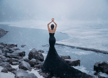 Sexy Woman Long Black Dress Silhouette Mermaid Train. Backdrop River Ice Water Stones Cold Blizzard Storm Snowfall. Lady Turned Away. Elegant Collected Hairstyle Bun. Bare Back Rear View Without Face