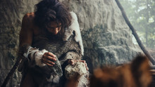 Close-up Shot Of A Primeval Caveman Wearing Animal Skin Hits Rock With Sharp Stone, Makes First Primitive Tool For Hunting Animal Prey. Neanderthal Using Flint Rock. Dawn Of Human Civilization.