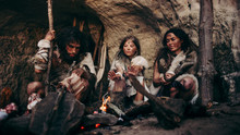 Tribe Of Prehistoric PrimitiveHunter-Gatherers Wearing Animal Skins Live In A Cave At Night. Neanderthal Or Homo Sapiens Family Trying To Get Warm At The Bonfire, Holding Hands Over Fire, Cooking Food