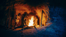 Tribe Of Prehistoric Hunter-Gatherers Wearing Animal Skins Stand Around Bonfire Outside Of Cave At Night. Portrait Of Neanderthal / Homo Sapiens Family Doing Pagan Religion Ritual Near Fire