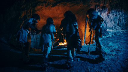 Tribe of Prehistoric Hunter-Gatherers Wearing Animal Skins Live in a Cave at Night. Neanderthal or Homo Sapiens Family Trying to Get Warm at the Bonfire, Holding Hands over Fire. Back View