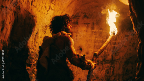 Portrait of Primeval Caveman Wearing Animal Skin Exploring Cave At Night, Holding Torch with Fire Looking at Drawings on the Walls at Night. Neanderthal Searching Safe Place to Spend the Night