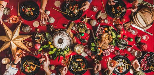 Wall Mural - Friends celebrating Christmas. Flat-lay of people eating and talking over festive table with red cloth with champagne, roasted chicken, bundt cake, fruits, decorations, top view, wide composition