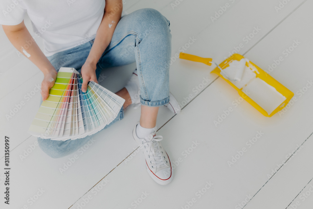 Faceless woman in jeans holds color samples, chooes best tone for refurbishment walls, sits on white floor, uses tray and paint roller for house remodeling. Home makeover and renovation concept - obrazy, fototapety, plakaty 