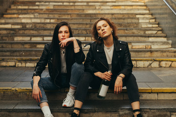 Wall Mural - Portrait of two girls models in stylish casual clothes sitting on stairs with bottle of wine in hand posing at camera with serious face. Two girlfriends are sitting on the stairs in the underground