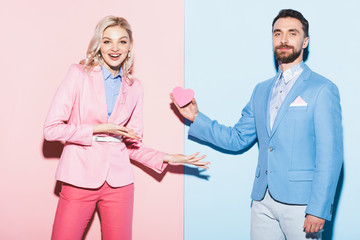 Wall Mural - handsome man giving heart-shaped card to shocked woman on pink and blue background