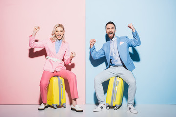 Wall Mural - smiling woman and handsome man sitting on travel bags and showing yes gesture on pink and blue background