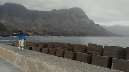 Wall Mural - Aerial view - a young woman tourist fooling around against the background of the ocean and the misty mountains. Agaete, Gran Canaria