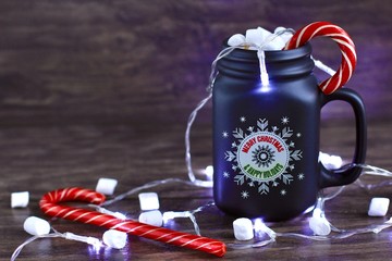 Mug of hot coffee with milk, marshmallow and Lollipop against rustic wooden background with Christmas decorations and lights.