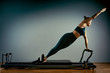 © Georgii - Young girl doing pilates exercises with a reformer bed. Beautiful slim fitness trainer on reformer gray background, low key, art light. Fitness concept