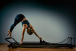 © Georgii - Young girl doing pilates exercises with a reformer bed. Beautiful slim fitness trainer on reformer gray background, low key, art light. Fitness concept