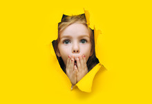 A Little Cute Girl With Red Hair Looks Out Through A Hole In Torn Yellow Paper And Covers Her Mouth With Her Hands And Eyes Wide In Surprise.The Concept Of Surprise, Shock. Discounts, Sales.Copy Space