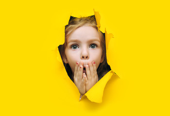 A little cute girl with red hair looks out through a hole in torn yellow paper and covers her mouth with her hands and eyes wide in surprise.The concept of surprise, shock. Discounts, sales.Copy space