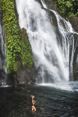 Wall Mural - Young slim brunette woman with curly hair enjoying in lagoon of huge tropical waterfall Banyumala in Bali. Wearing in black swimsuit. Happy vacations in Indonesia. Wanderlust travel concept.