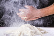 Close up of chef human's hands preparing for kneading the dough on the table, powdering with flour. Making dough by hands at bakery or at home. Flour cloud in the air. Black background.