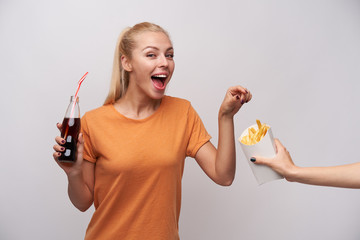 Wall Mural - Joyful young attractive blonde female with ponytail hairstyle keeping bottle with straw in raised hand and reaching for french fries with wide happy smile, isolated over white background