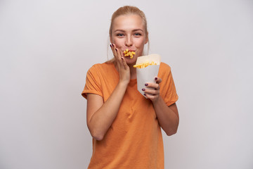 Wall Mural - Shot of positive young pretty blonde female with casual hairstyle looking cheerfully at camera with mouth full of french fries, being hungry and happy to get meal, isolated over white background