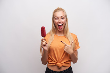 Wall Mural - Overjoyed lovely young blonde woman with casual hairstyle pointing with forefinger on berry ice-cream in her hand, looking joyfully to camera with wide mouth opened, isolated over white background